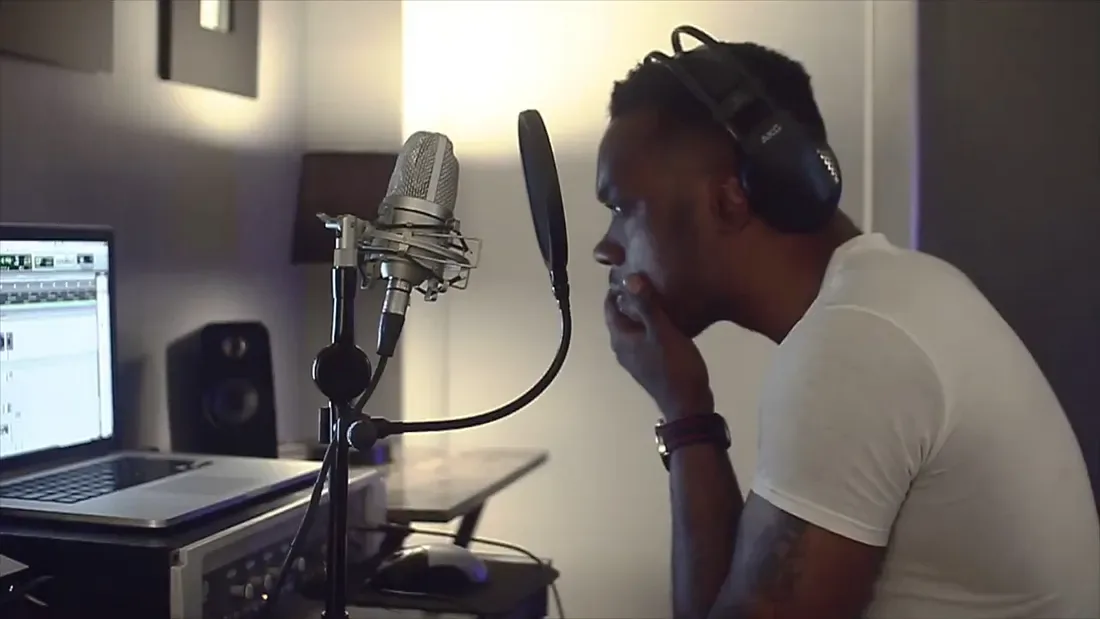 A singer in headphones at a home-studio microphone, with a laptop showing a digital audio workstation and a studio monitor on the desk